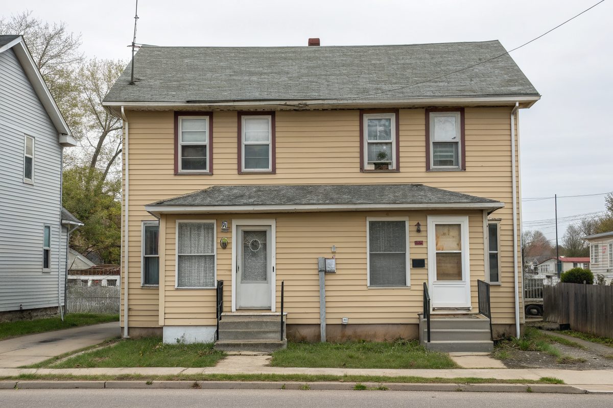 An older two-story duplex with tan siding and a worn gray shingle roof